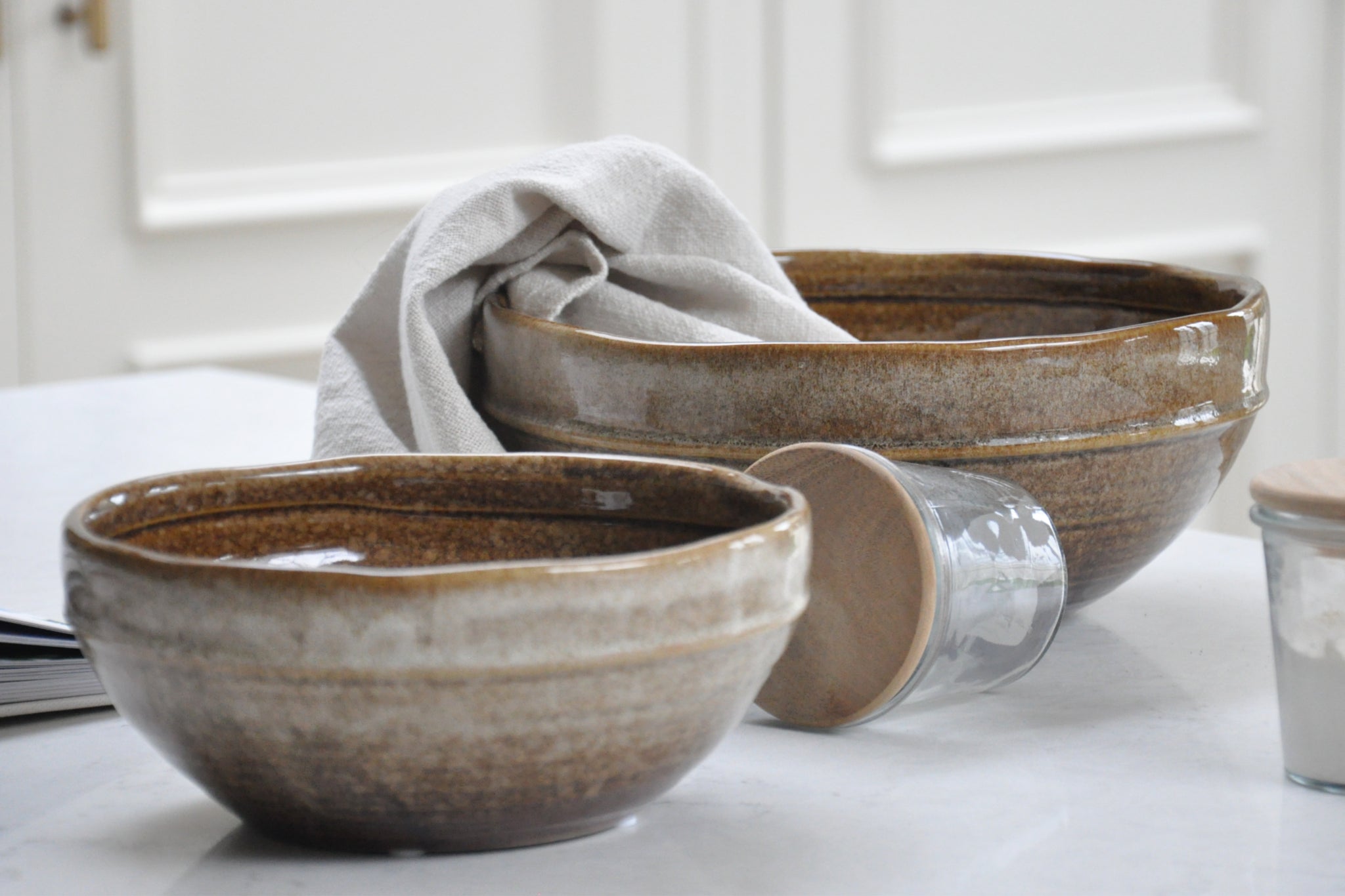 Kitchen still of brown bowls, beige tea towel, and rustic storage jars scattered on a granite countertop